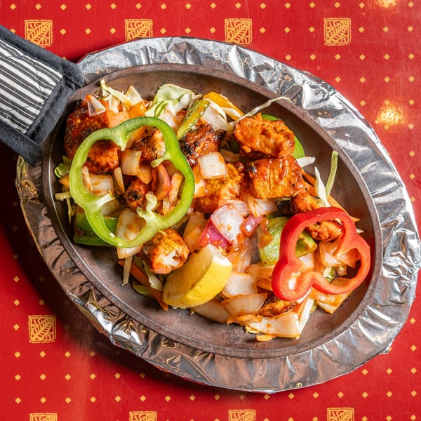 Sizzling chicken tikka with bell peppers, onions, and lemon wedges on a red patterned tablecloth.