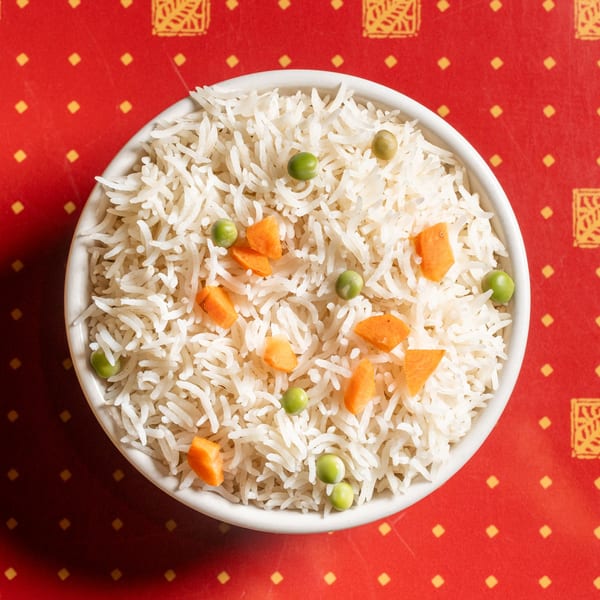 White rice with green peas and diced carrots in a bowl on a red patterned surface.