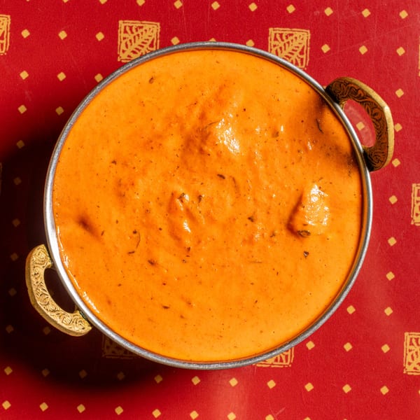 Top-down view of a metal bowl filled with vibrant orange butter chicken on a red patterned tablecloth.