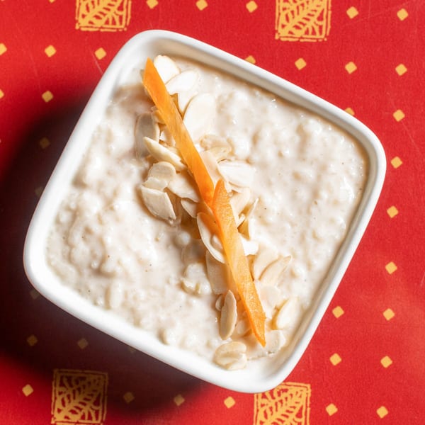 Creamy rice pudding topped with sliced almonds and an orange peel, on a red patterned tablecloth.